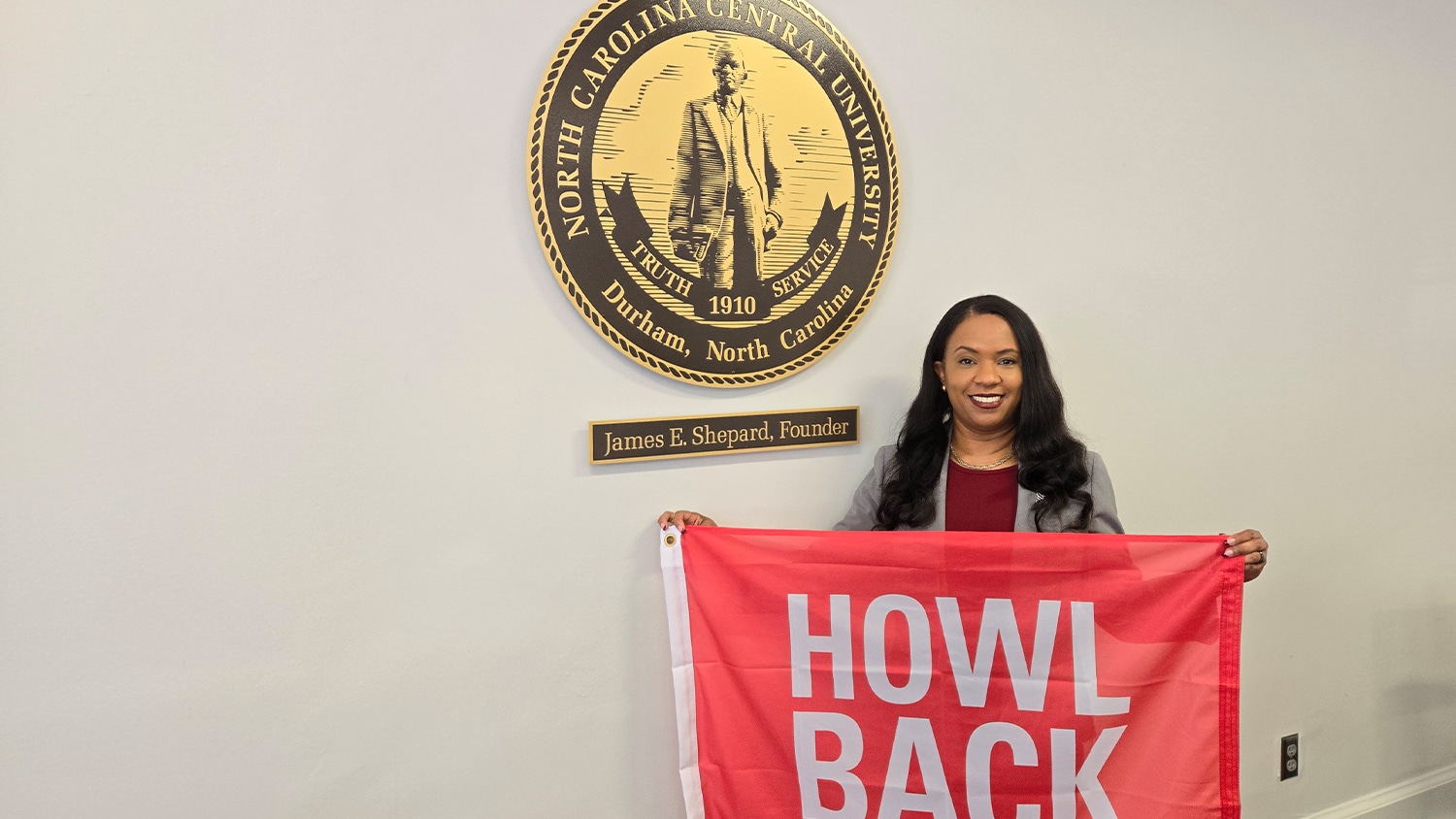 North Carolina Central University Chancellor Karrie Dixon holds a red banner than reads "Howl Back" while standing in front of the seal for North Carolina Central University on a white wall.