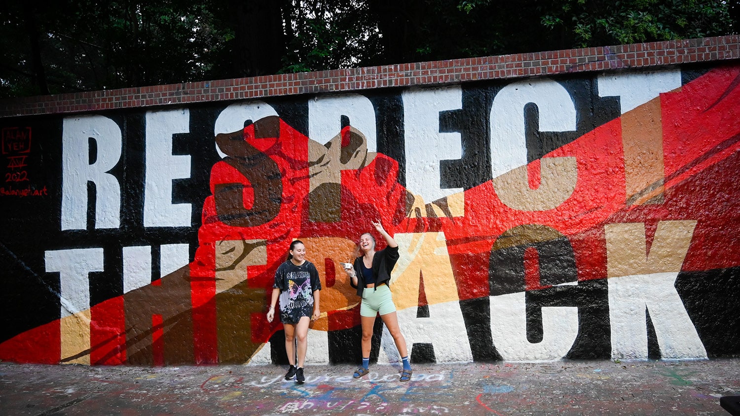 Two students laugh together while standing in front of a mural that shows two hands clasped together and reads "Respect the Pack".