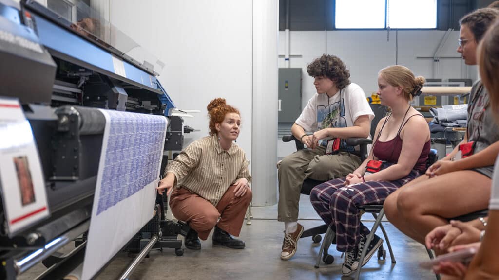 Bailey Knight crouches in front of a fabric printer while a piece of blue and white fabric is being printed. She speaks with high school students, who watch the printing process.