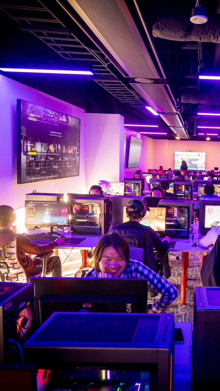 A student smiles while looking at a computer screen. In the background, other students sit at rows of computers playing esports games.