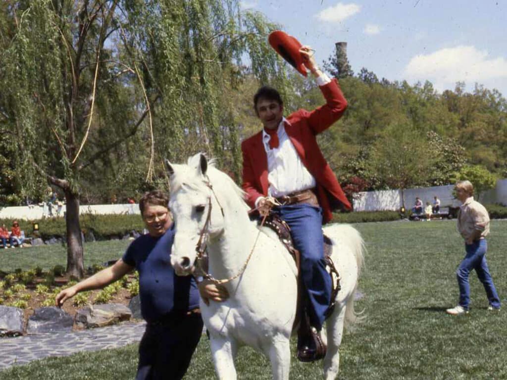 Kiffin, wearing a red cowboy outfit complete with 10-gallon hat, rides a white horse on the grounds of NC State's campus