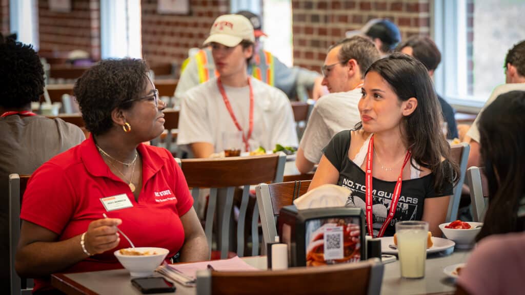 Wearing a red NC State polo, Jada Brewington speaks with Medisa Elyasi at a dining hall table. They both have bowls of food in front of them.