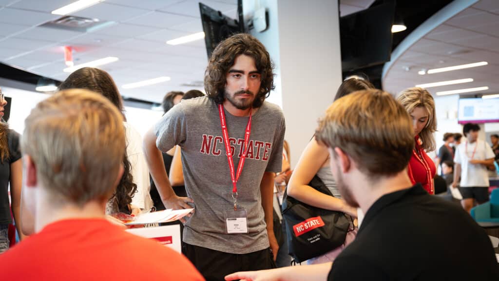 A male student wearing an NC State t-shirt and lanyard talk with two students sitting at a table during an information fair.