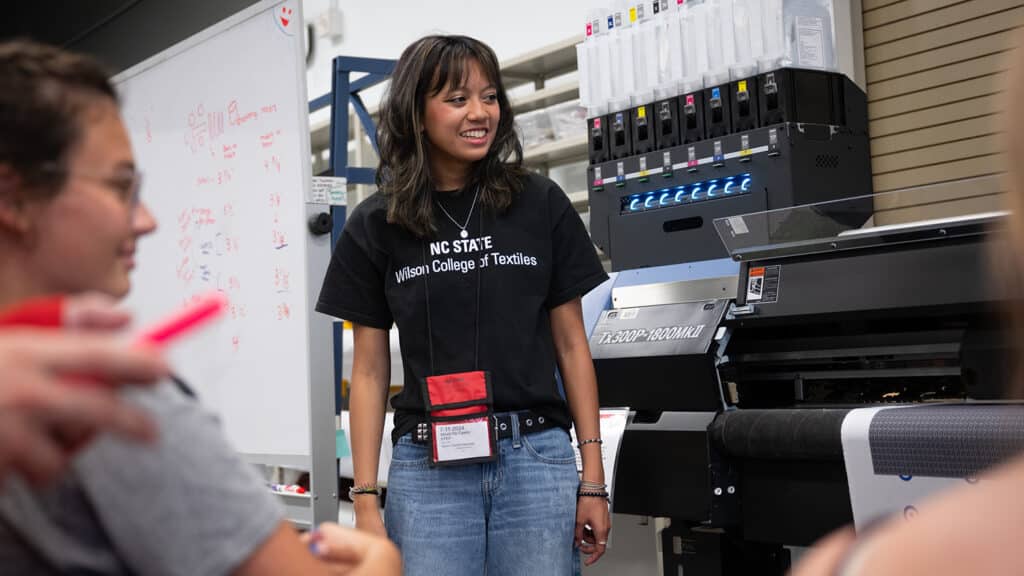 NC State sophomore Alexis De Castro stands in front of a fabric printer and talks with high school seniors, who are sitting in front of her. De Castro wears a black Wilson College of Textiles T-shirt. 