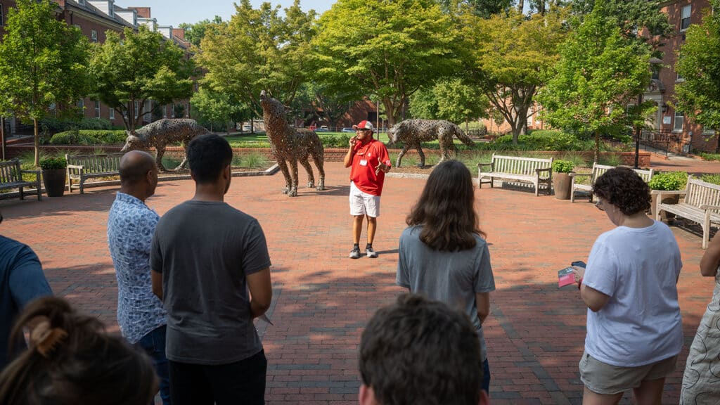 Travis Espinoza wears a red NC State polo and baseball cap as he speaks to a small crowd of campus visitors. They look at the three bronze wolf sculptures that stand in the background. 