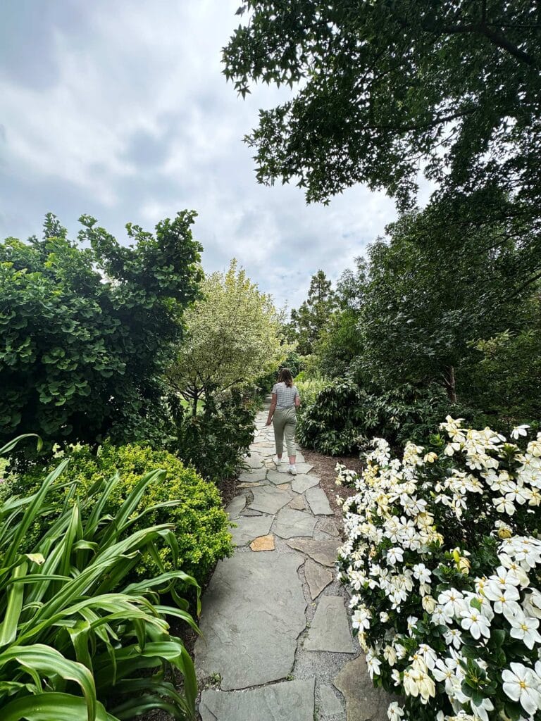 A woman walks down a stone path running between bushes, trees and flowers in a garden.