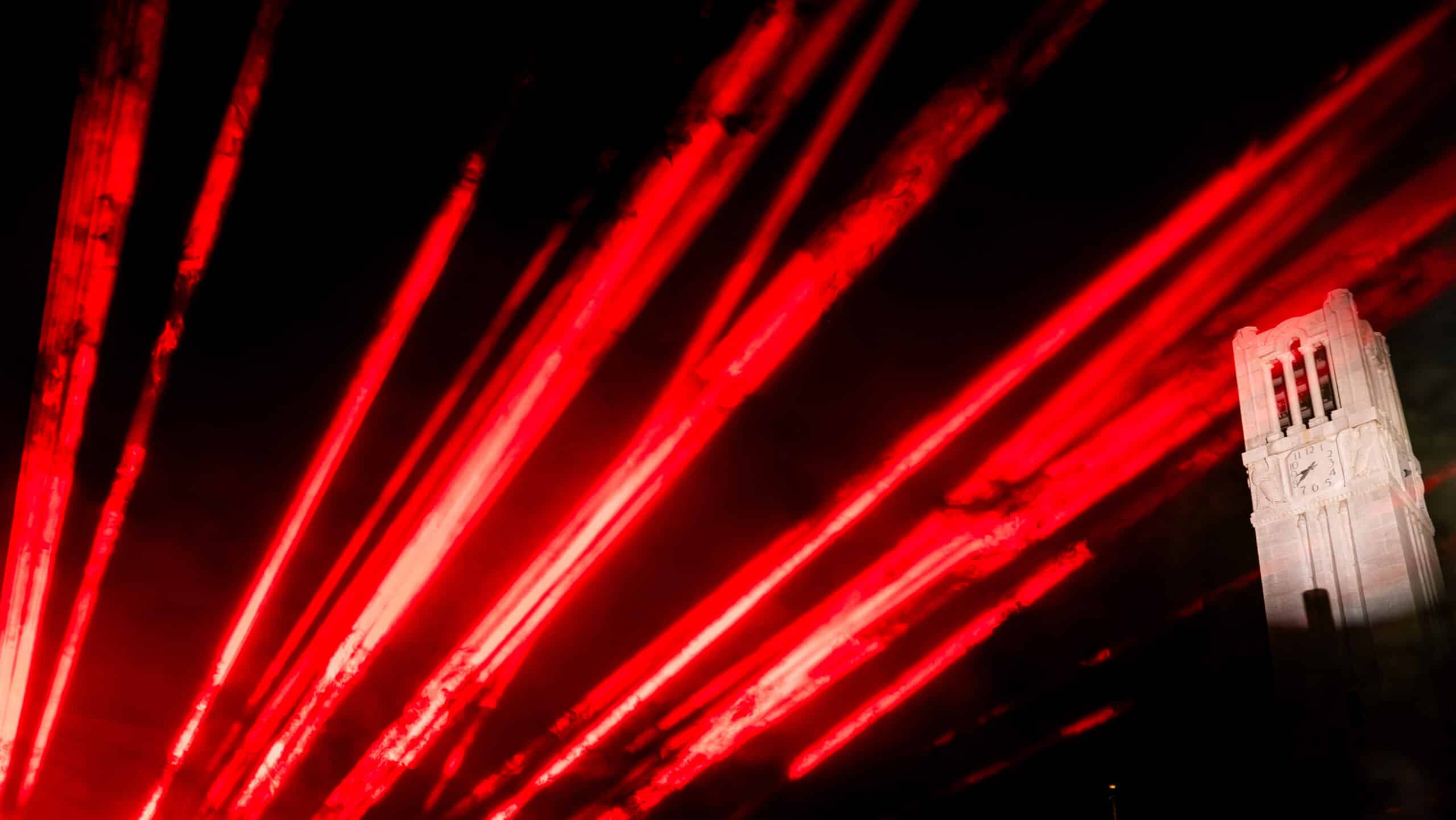 The NC State Belltower at night with red lights streaking toward it.