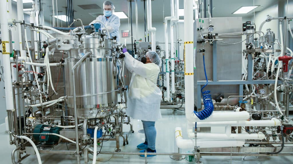 A person in a lab coat, face mask and hair net checks a metal container in a biomedical facility. 