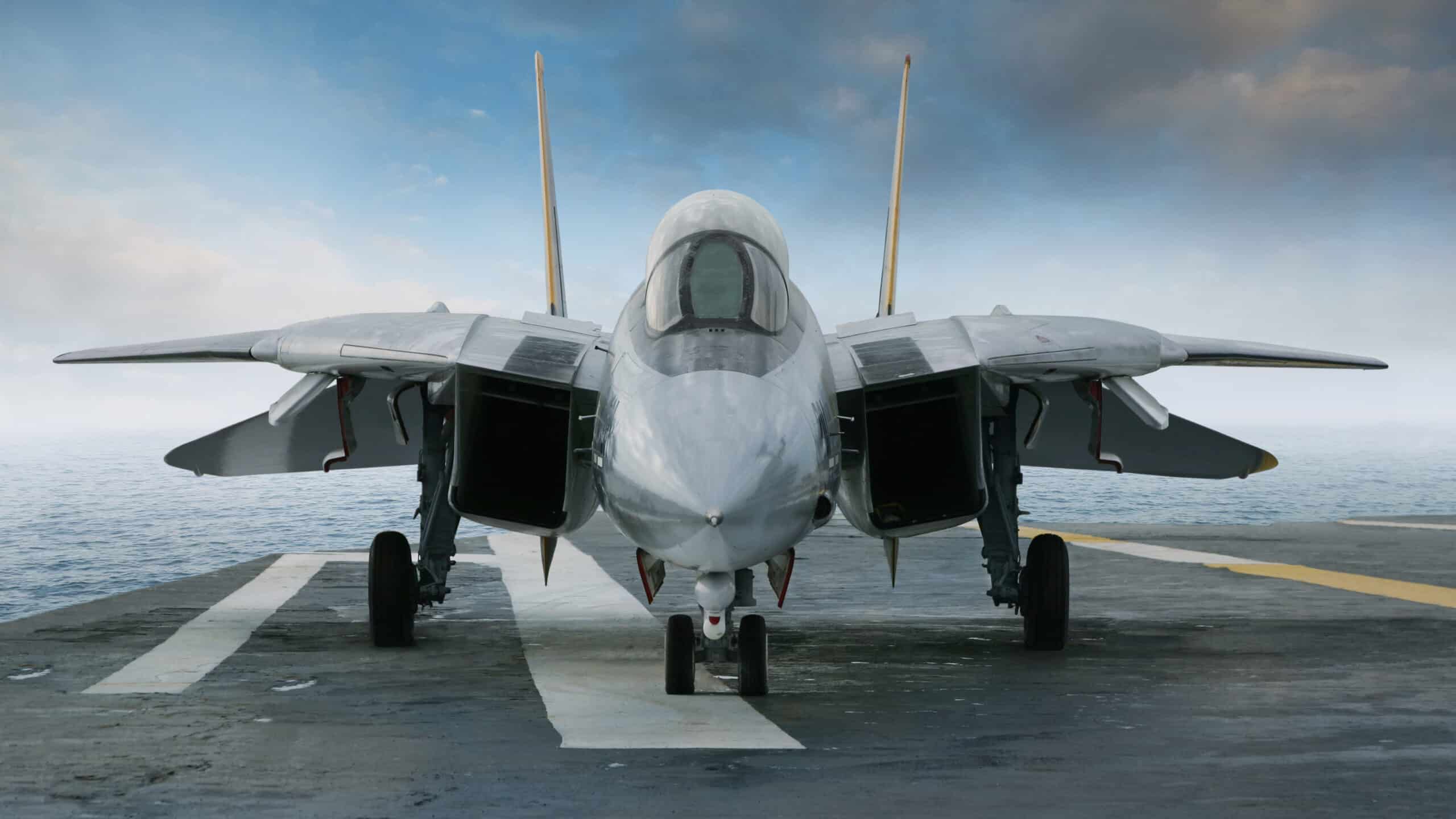 An F-14 jet fighter on an aircraft carrier deck beneath blue sky and clouds viewed from front.