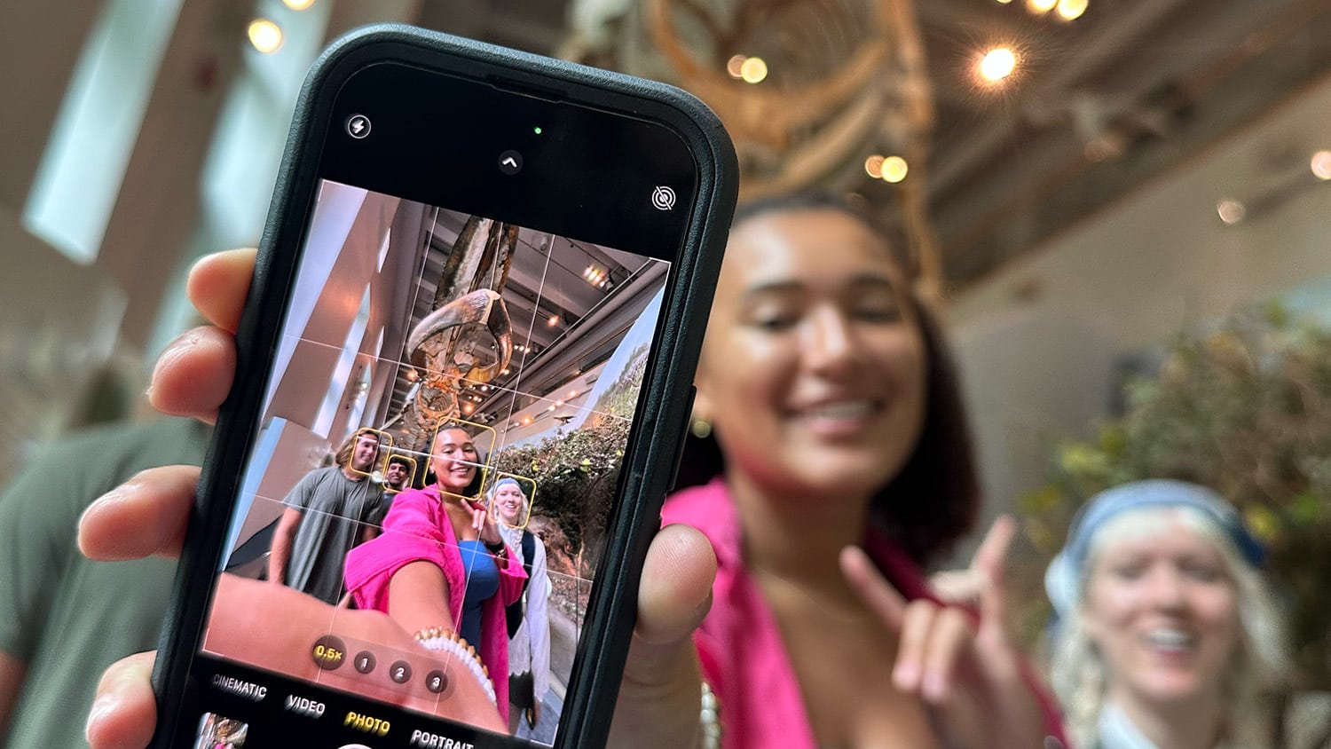 A close-up of the screen of a cell phone which is pointed toward, and displaying, a group of students standing in front of a large skeleton of a whale.