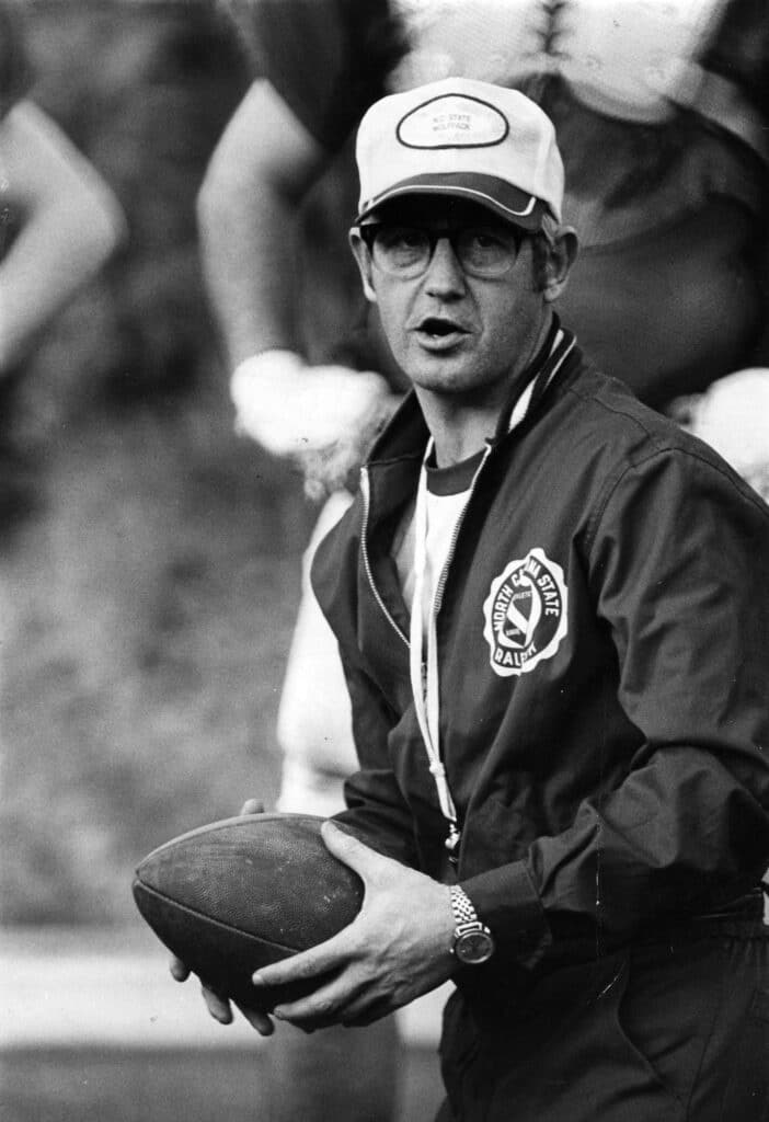 Coach Lou Holtz wearing an NC State jacket and a baseball cap and holding a football