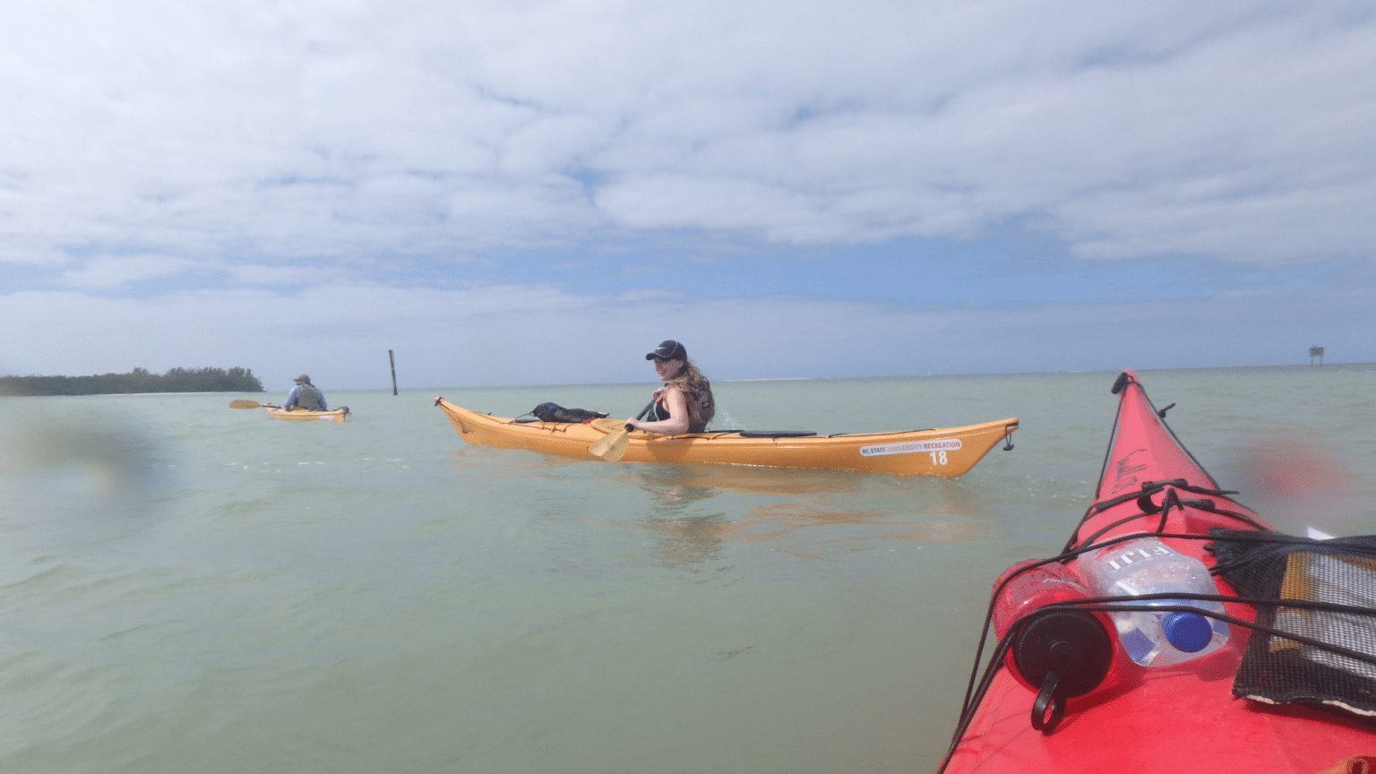 Seen from the point of view of a person in a kayak, two other students row kayaks across a body of water.