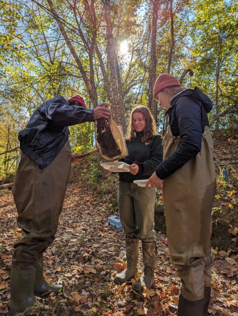 A photograph of three students wearing waterproof clothing inspect samples in the woods.