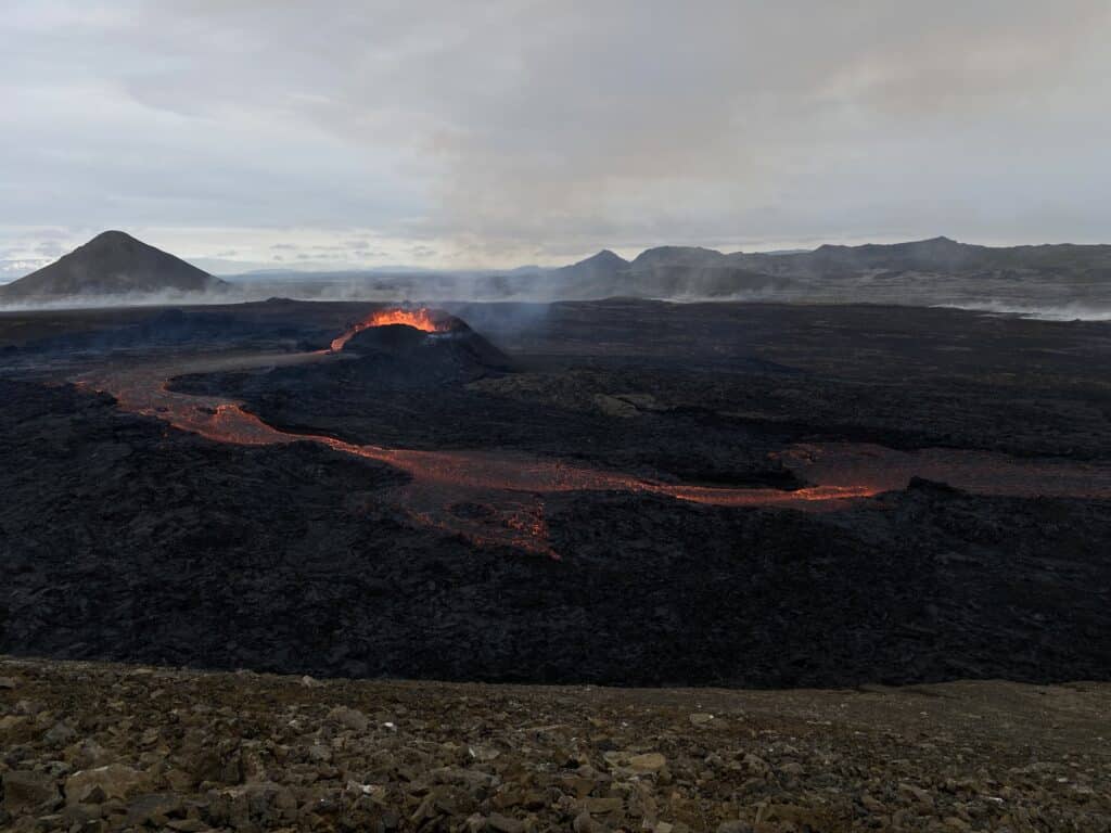 A photograph of a volcano erupts with a flow of bright orange lava in an area surrounded by blackened earth and mountains in Iceland.