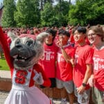 A view of Ms. Wuf holding a cellphone and taking a selfie, with four NC State students standing directly behind her and a larger crowd of students in the background.