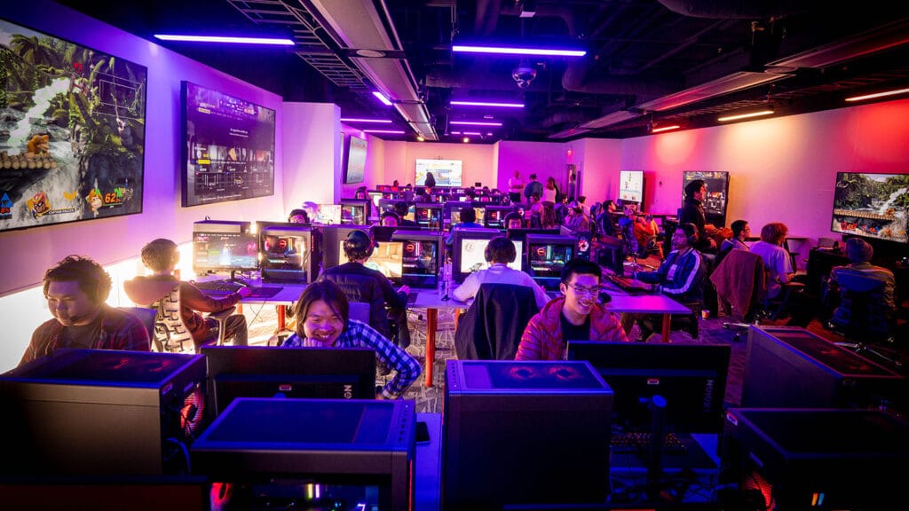 A view of the Gaming and Esports Lab at the Hunt Library, with students gaming at various stations, dimmed ambient lighting and neon blue and orange lighting illuminating the scene.