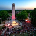 An overhead shot of the Belltower lighted red at dusk, with a crowd of NC State supporters gathered around it in the foreground.