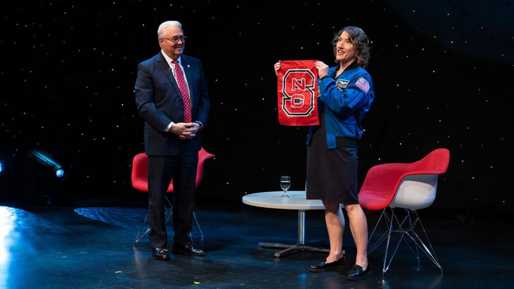 Chancellor Randy Woodson and three-time NC State alum and NASA astronaut Christina Koch stand on a stage backlit by stars. Koch holds an NC State flag up to view for an unseen crowd of NC State supporters.