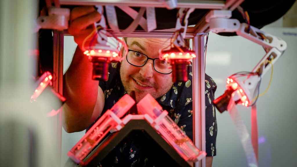 A shot of a researcher's face from the other side of a contraption that the researcher is looking into while also reaching in to manipulate something in the device.