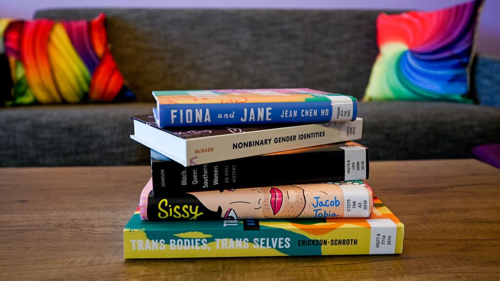 Several books sit atop a wooden table in the LGBTQ Pride Center, with a couch and multi-colored couch pillows out of focus in the background.