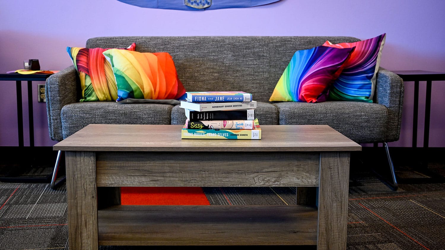 Several books sit atop a wooden table in the LGBTQ Pride Center, with a couch and multi-colored couch pillows in the background.