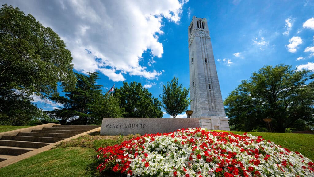 An upward looking shot of the Belltower with a partly cloudy blue sky behind it, and red and white flowers blooming in front of it in the foreground. Green-leafed trees also feature prominently in the middle ground near the Belltower.