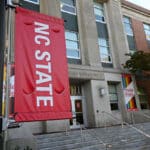 A view of an NC State flag hanging outside the Poole College of Management. A rainbow-colored Poole Pride flag hangs on the building in the background.