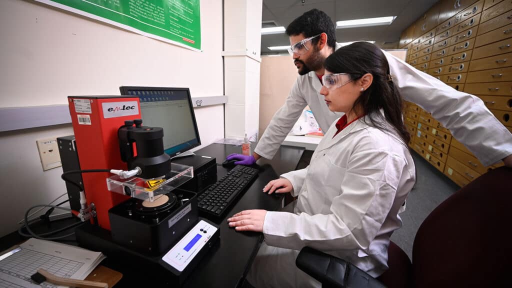 Two researchers wearing lab coats and goggles, one sitting and one standing, gaze at a computer screen.