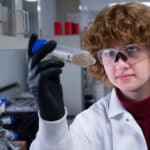 A close up shot of a researcher holding a test tube up and peering at its contents, which appear to be a liquid clouded with substrate. The researcher wears a lab coat and protective goggles as she stands in a laboratory, with a rounded device for holding more test tubes appearing behind her in the bottom left.