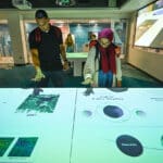 Two people stand looking down at a table featuring a digital visualization.