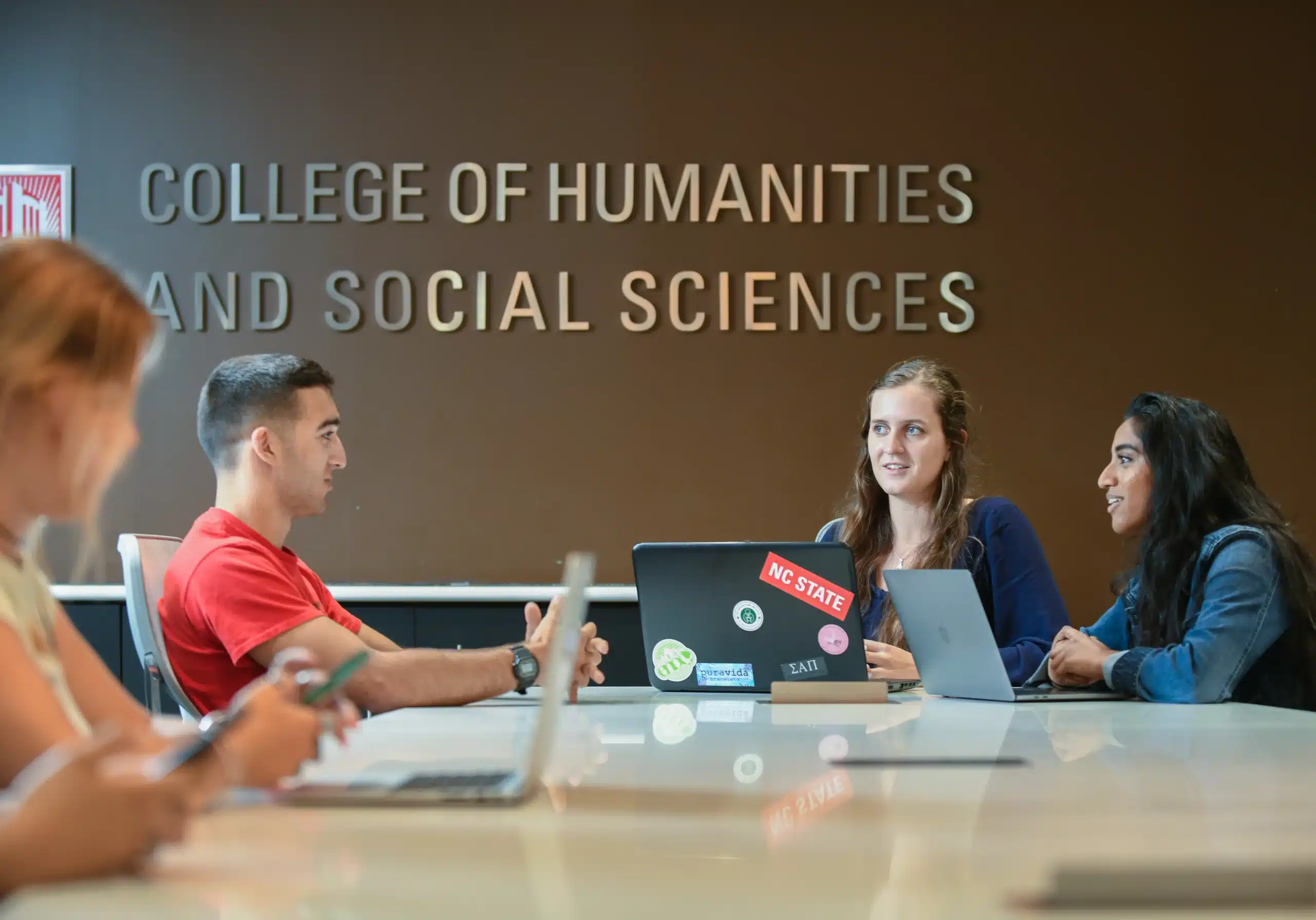 Students sit at a large table in front of the sign for the College of Humanities and Social Sciences.