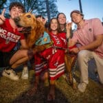 A group of NC State supporters pose with their dog, a Golden Retriever, who's adorned in a Wolfpack athletics jersey. The supporters are showing the Wolf Ears hand signal.