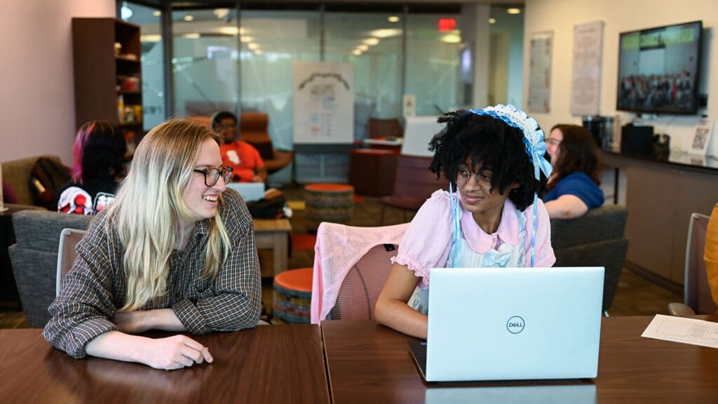 Two students smile while sitting together at a table in the LGBTQ Pride Center, while