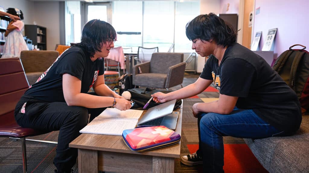 Two students study together at a table in the LGBTQ Pride Center. One student sits in a chair, the other on a couch. One student points out something in a notebook with a pen in hand.