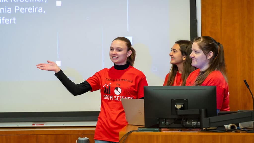 Three students make a presentation in front of a screen while standing behind a podium and smiling at an unseen audience. They wear Wolfpack Red shirts that say "Easily distracted by Data Science."