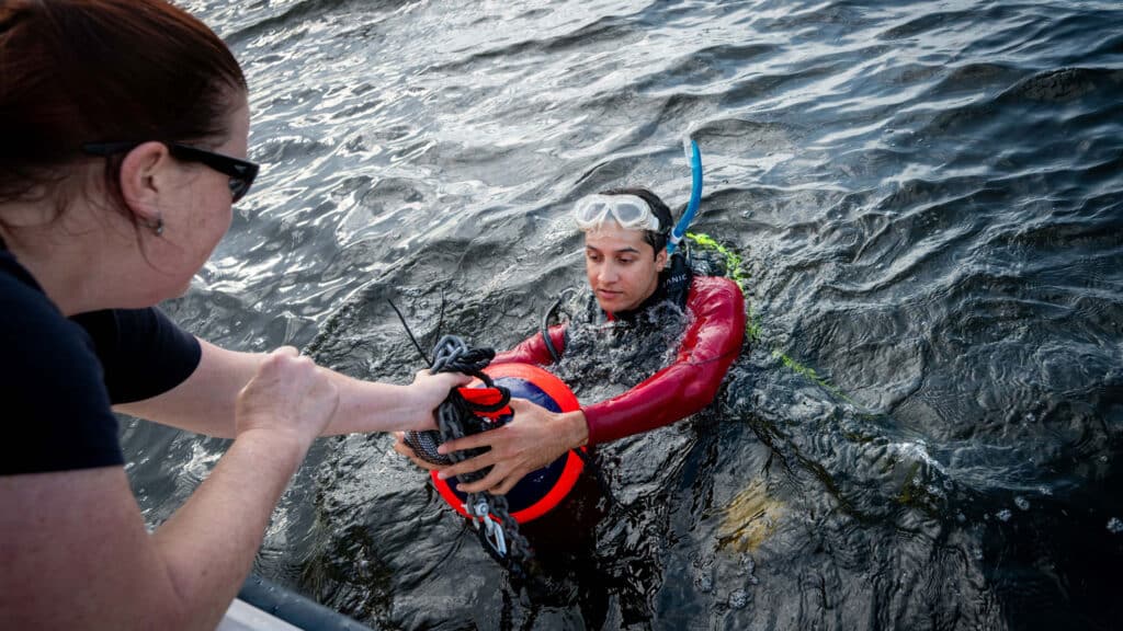 Astrid Schnetzer leans down from a boat to hand Zakir Bulmer, who is in the water wearing diving gear, some materials.