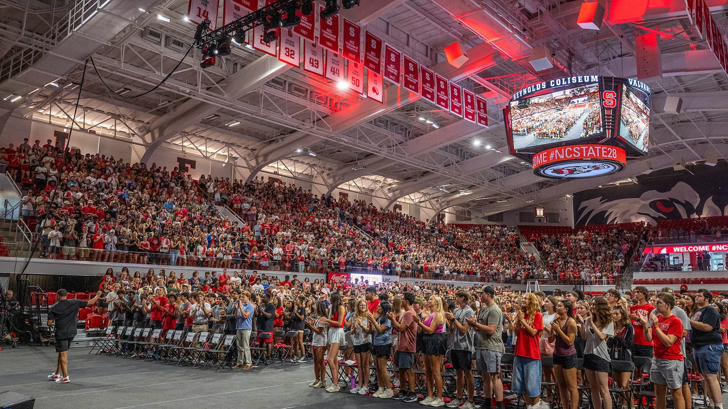 A large crowd of students stands on the floor and in the stands of Reynolds Coliseum during New Student Convocation.