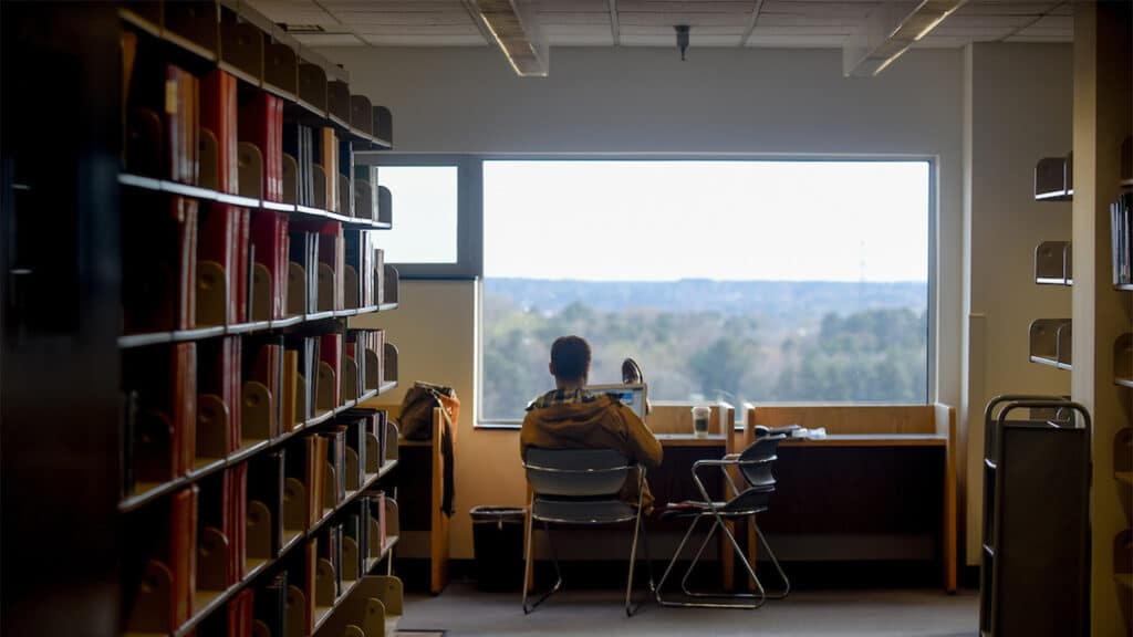 A student sits by a large window between the book stacks in Hill Library