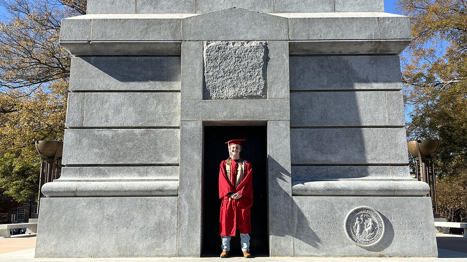 An NC State graduate poses in his red regalia while standing in the doorway of the Memorial Belltower.