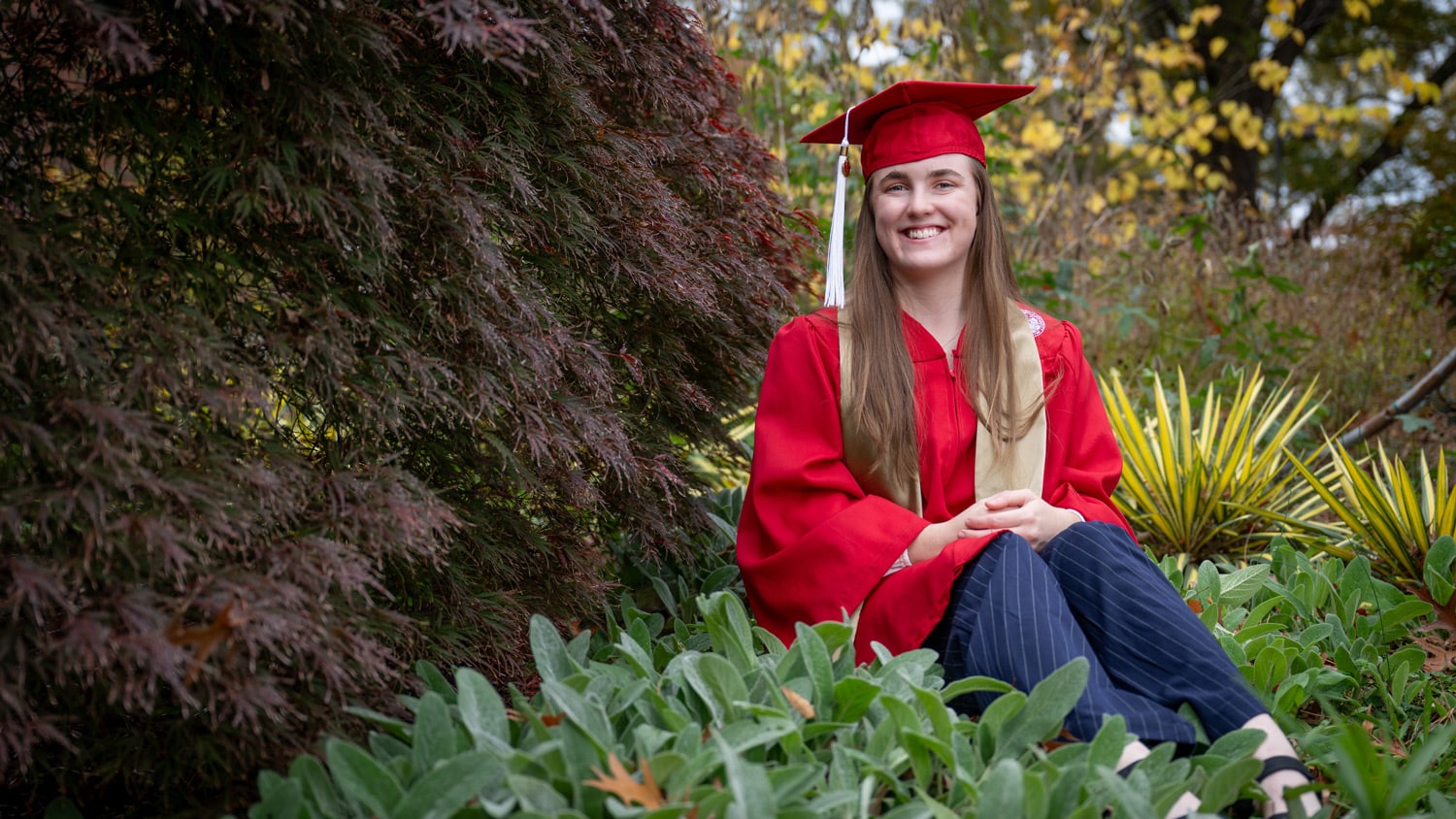 Lauren Turner sits by a tree in her red cap and gown