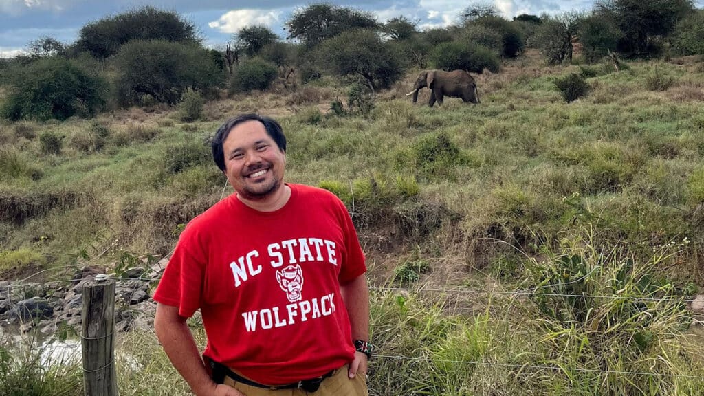 Matthew Snider smiles at the camera as an elephant walks by behind him