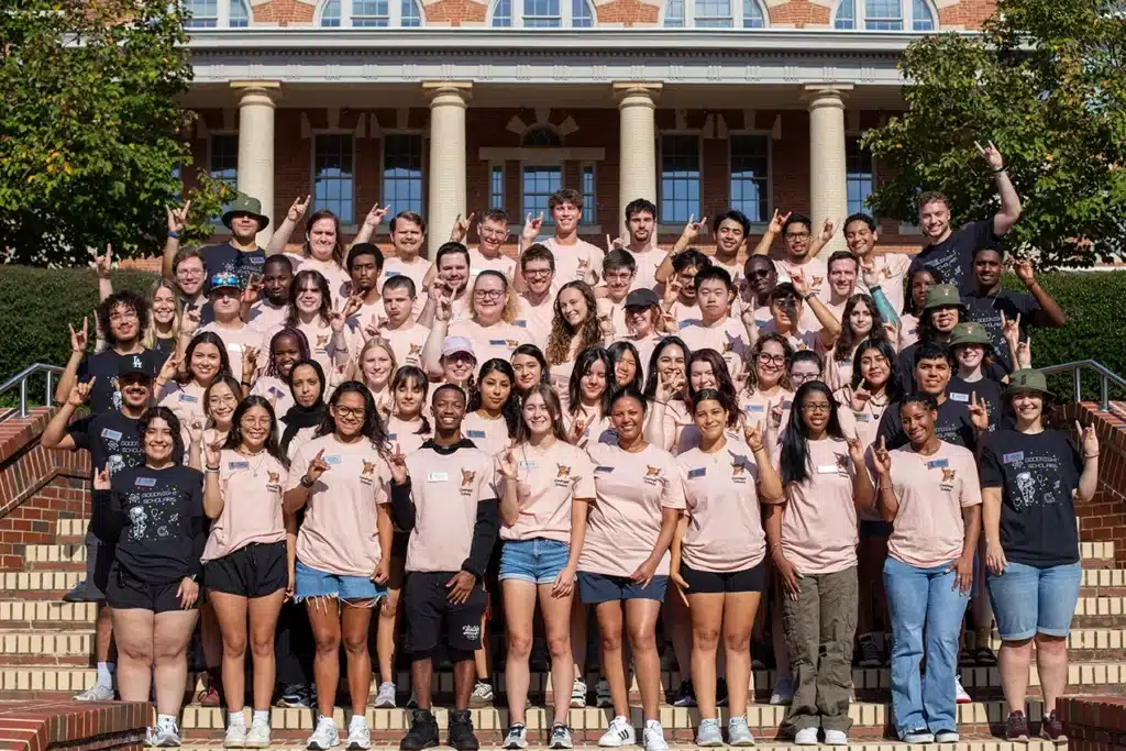 A large group of Goodnight scholars make the Wolfpack sign standing on a building's steps. 