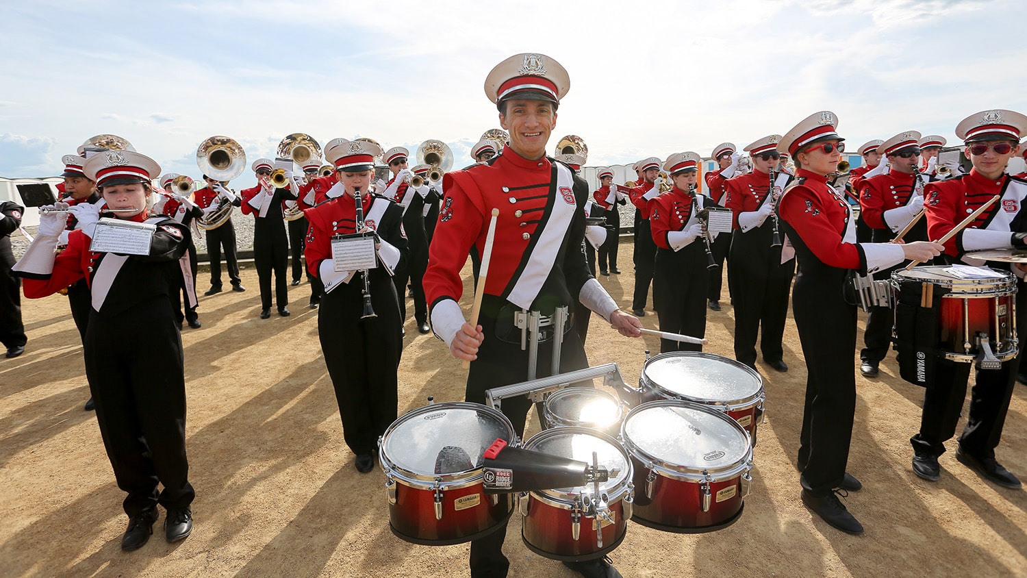 A student in a marching band uniform plays the quad drums surrounded by other members of the band on a beach.
