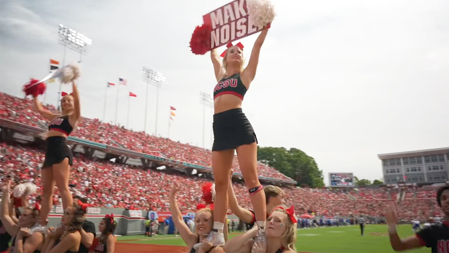 Two cheerleaders hold up signs while being supported in the air by more cheerleaders, on the field at Carter-Finley Stadium.