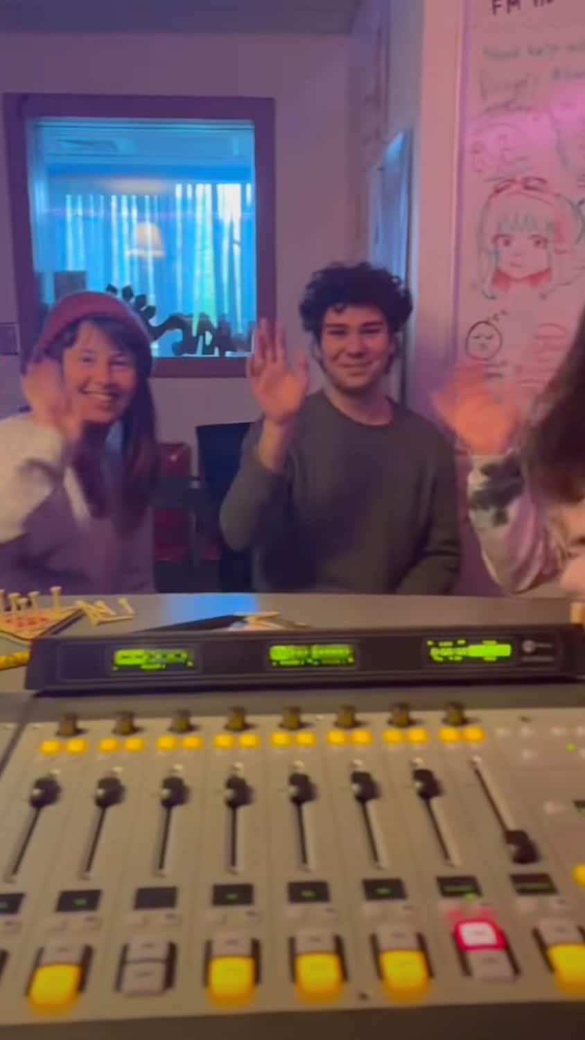 Two students wave from behind a sound board in a radio studio.