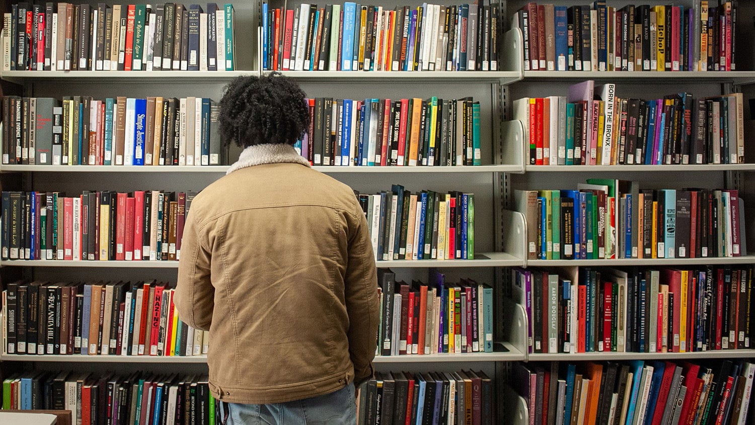 An NC State student stands with their back to the camera facing shelves filled with books.