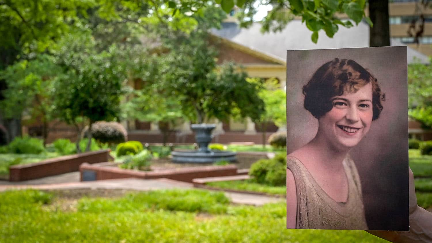 A photo of a young Mary Yarbrough, the first woman to earn a graduate degree from NC State, is held up to the camera, with the greenery of Mary Yarbrough Court showing in the background.