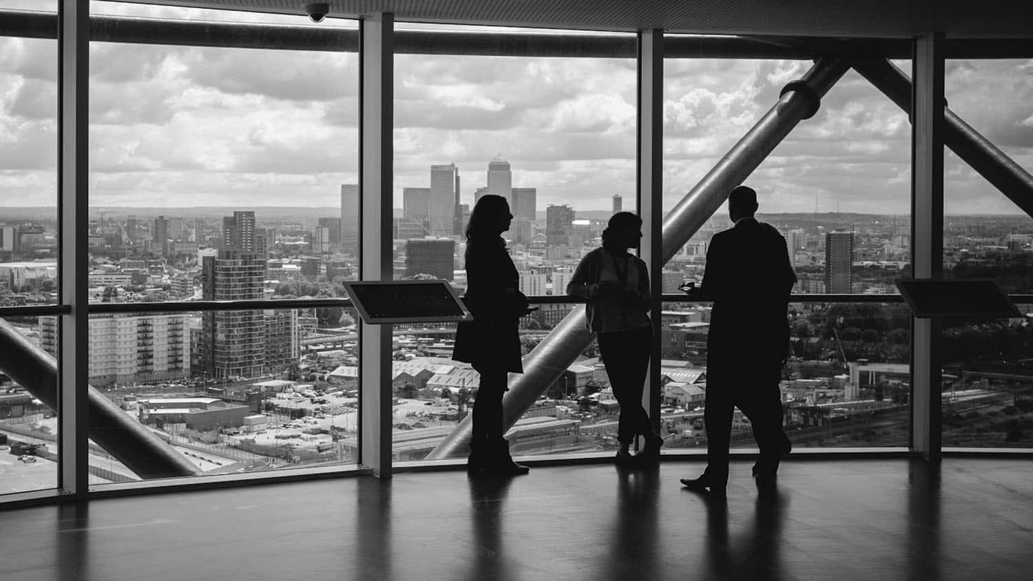 a man in a suit stands in front of a window talking to two other people