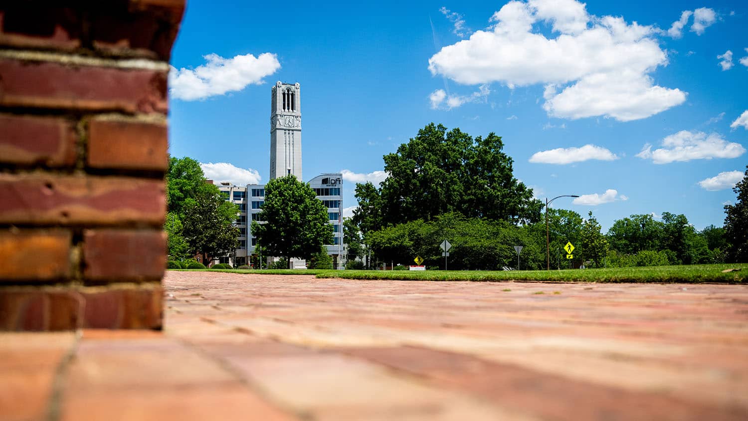 A brick pathway leads toward a tall clock tower under a bright blue sky with fluffy clouds, framed by a brick wall on the left.